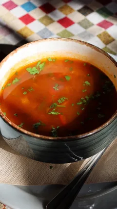 Un caldo rojizo con trozos de verdura y perejil picado por encima, en un cuenco de cerámica oscura.